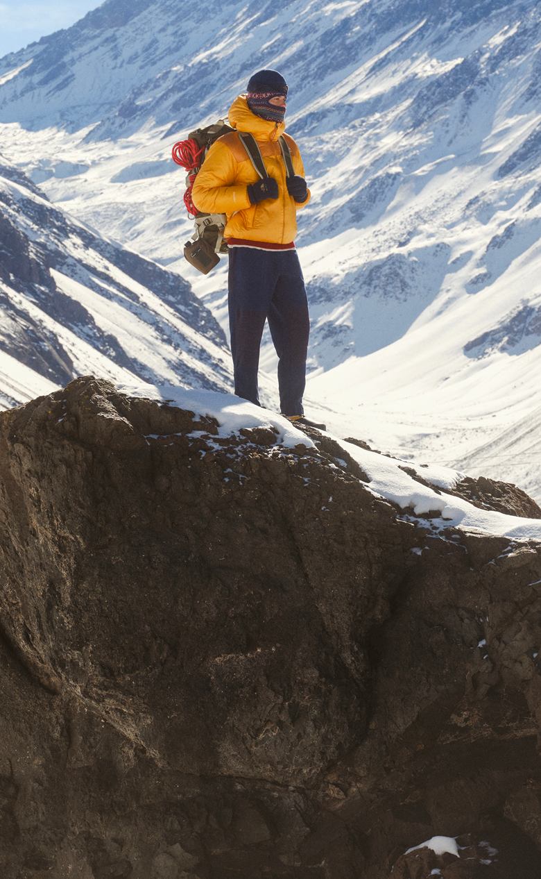 Photo plein pied d’un homme vêtu d’un manteau jaune et d’un pantalon marine, tenant un sac à dos, debout devant une montagne enneigée.  