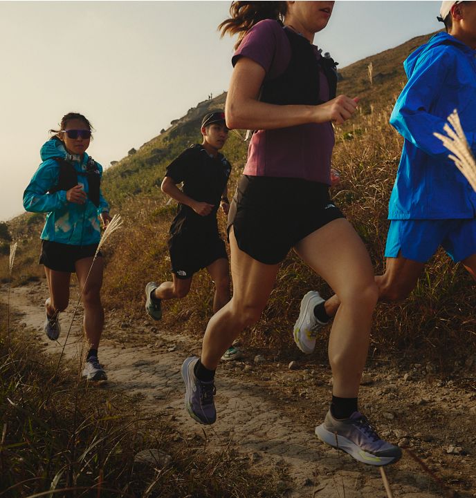 A group of four runners running uphill in soft lighting. The woman leading the way is in a blue jacket, black running shorts and purple/pink lensed sunglasses.