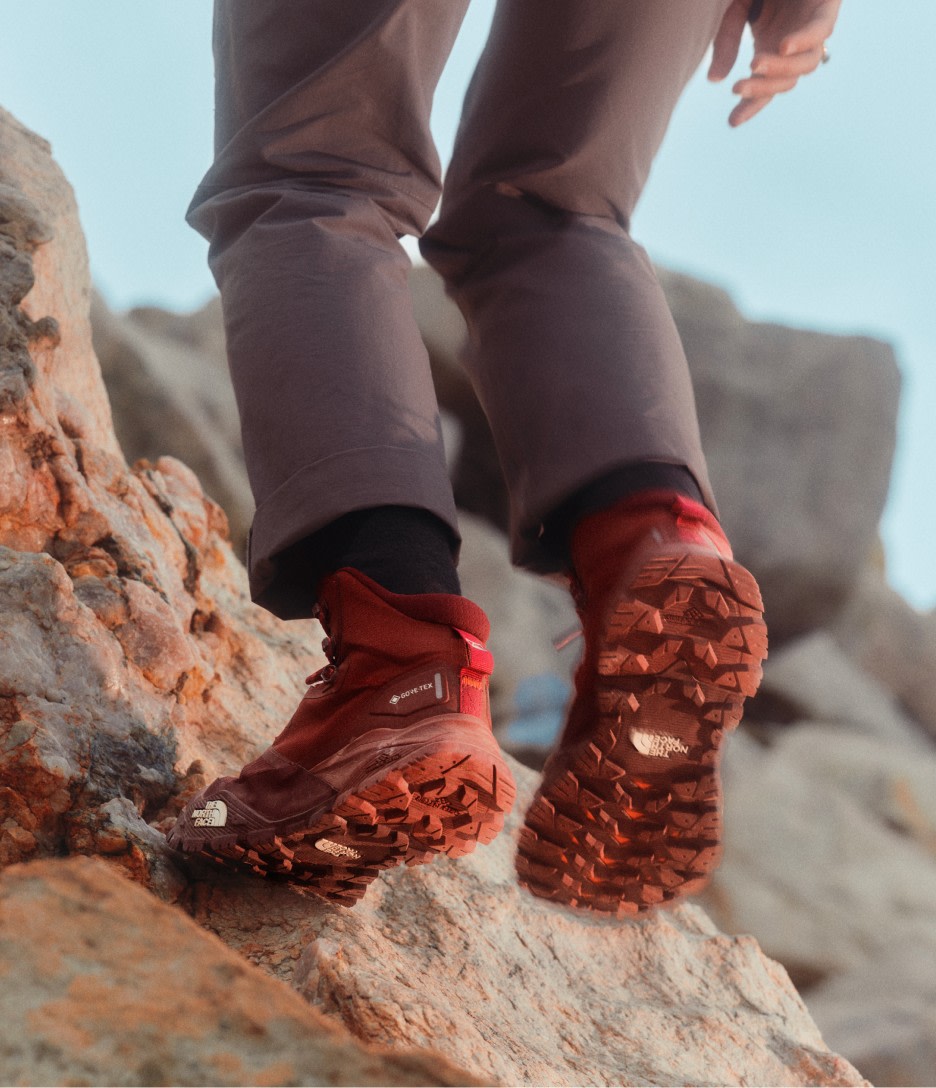 A woman stepping up onto a rock wearing red hiking boots and gray pants.